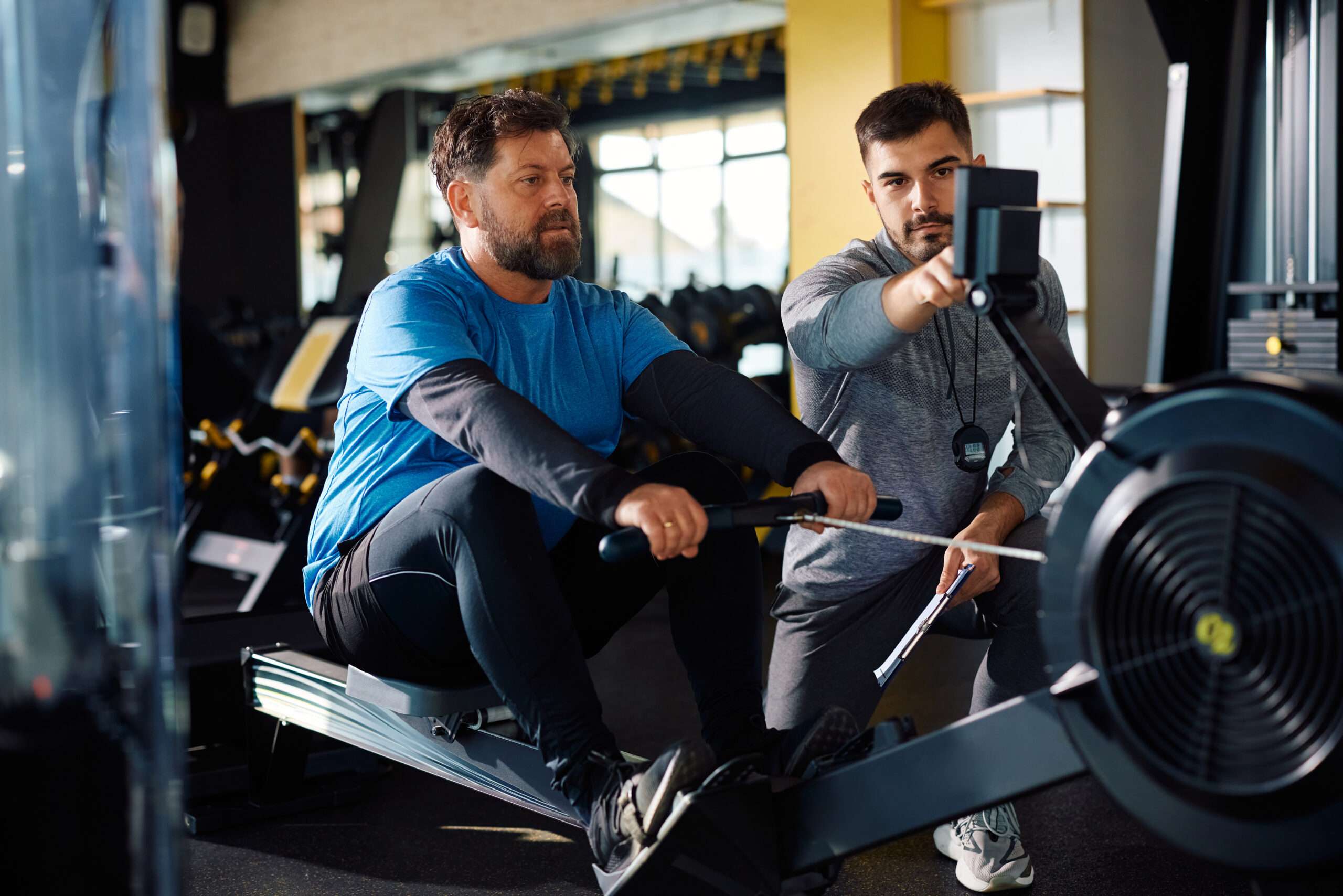 Mature man exercising on rowing machine with coach's assistance in fitness center.